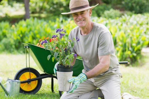 A cheerful middle-aged man wearing a light grey t-shirt, beige trousers, a straw hat, and gardening gloves is kneeling on a well-maintained lawn in a garden. He is holding a small potted plant with purple and red flowers, which he appears to have just picked or prepared for planting. Behind him, a bright yellow wheelbarrow with black handles and wheel is positioned on the grass, containing a few gardening tools and a green plant pot. The garden features a lush, neatly trimmed lawn with vibrant green grass, surrounded by dense foliage, including a hedge in the background and several tall trees that provide dappled sunlight. The scene suggests a pleasant day with natural light and clear weather, emphasizing a peaceful outdoor setting suitable for gardening services such as lawn and garden maintenance by Lawn Mowing Seven Sisters, serving the local area near SEvens Sisters in the UK.