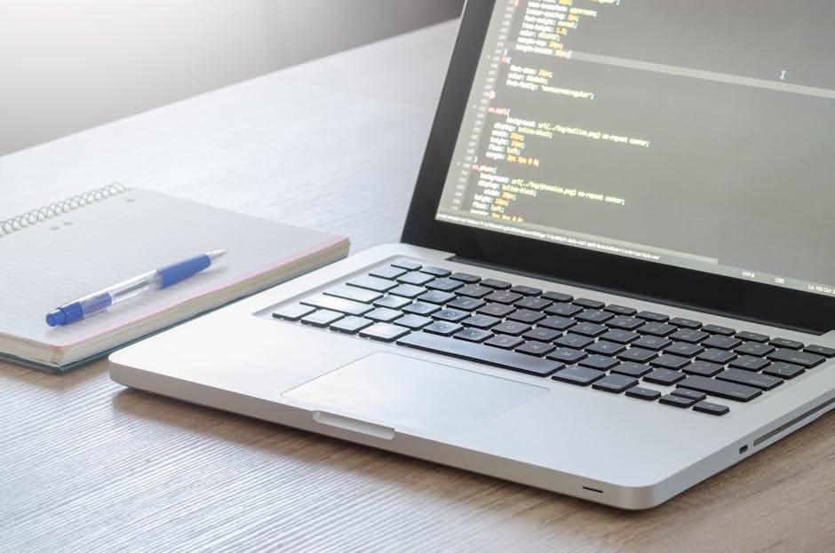 A close-up view of a silver laptop with a black keyboard displaying lines of HTML code on its screen, placed on a light wooden desk. To the left of the laptop, there is a spiral-bound notepad with fai