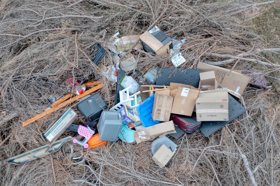 A ground-level view of a vacant outdoor area featuring a pile of discarded household and garden waste, including various cardboard boxes, plastic containers, a small plastic lawn chair, a metal storag